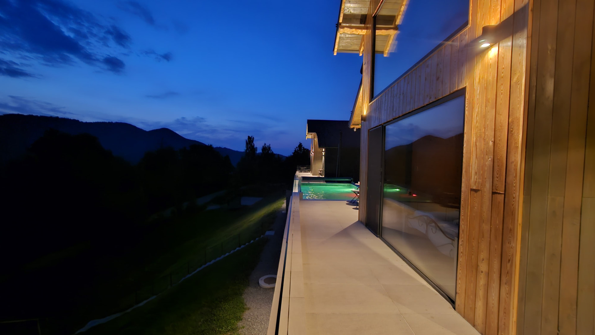 The same terrace in the blue of evening, the pool water lit green and the façade lamp glowing