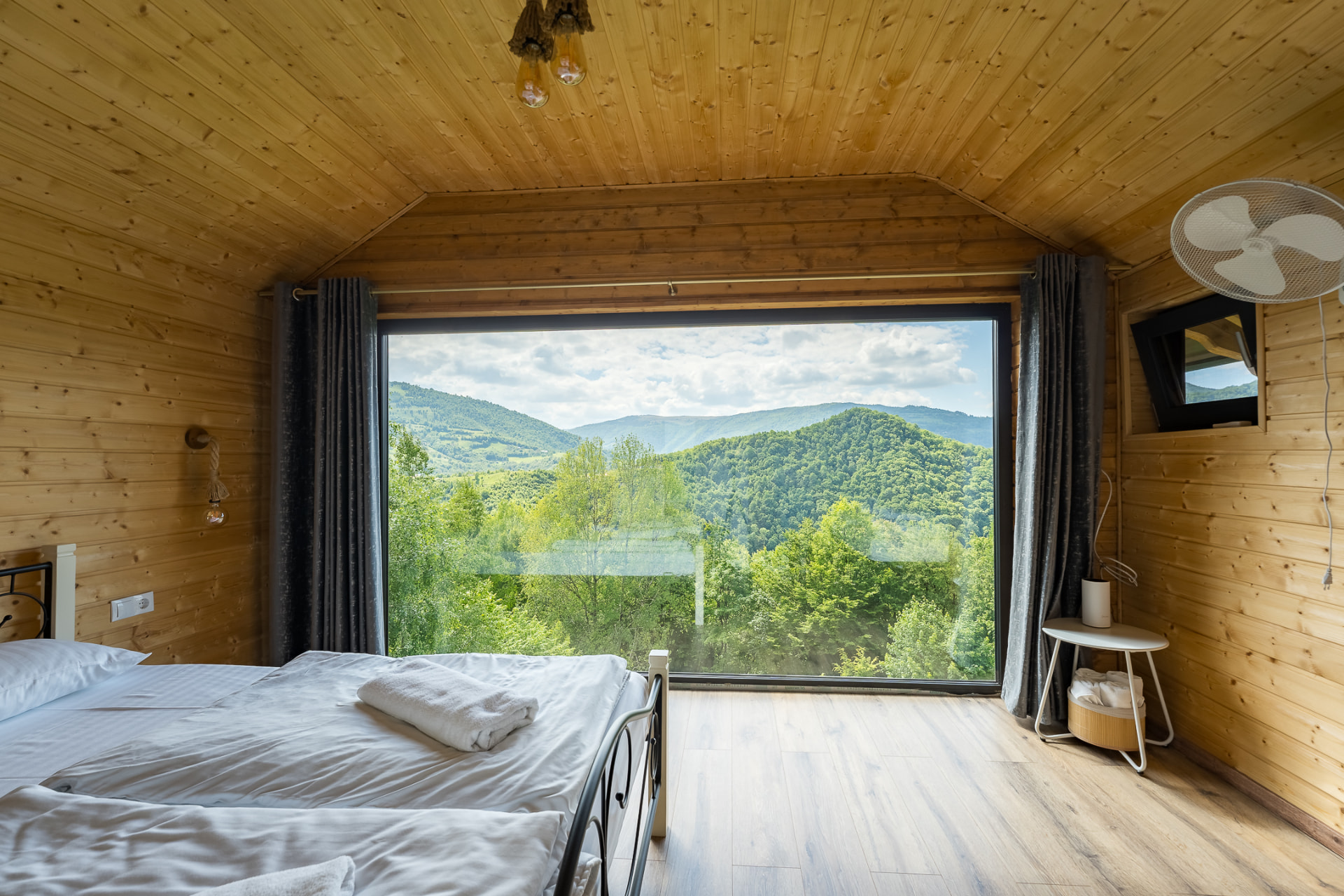 Dormitor din lemn cu pat dublu și fereastră panoramică ce dă spre dealurile împădurite