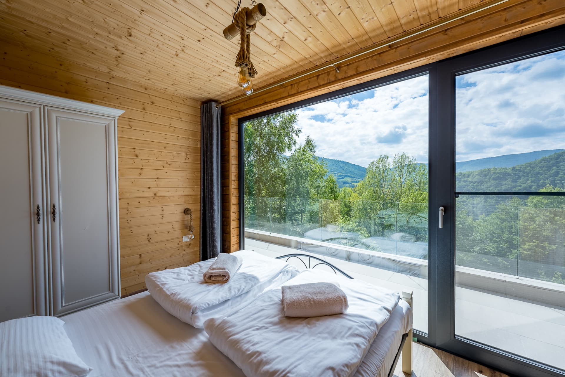 Wooden bedroom at Cabanele Carmel, with a double bed and a large window facing the forested hills