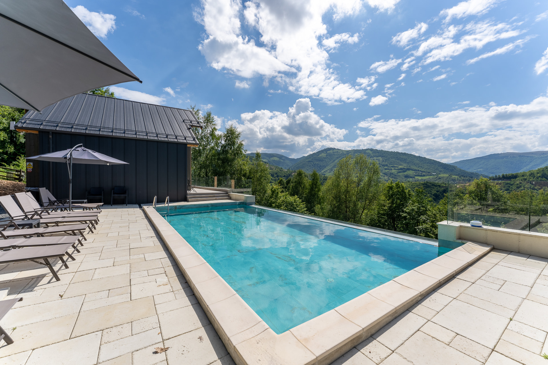 The outdoor pool with turquoise water, loungers and umbrellas, above a valley of forested hills