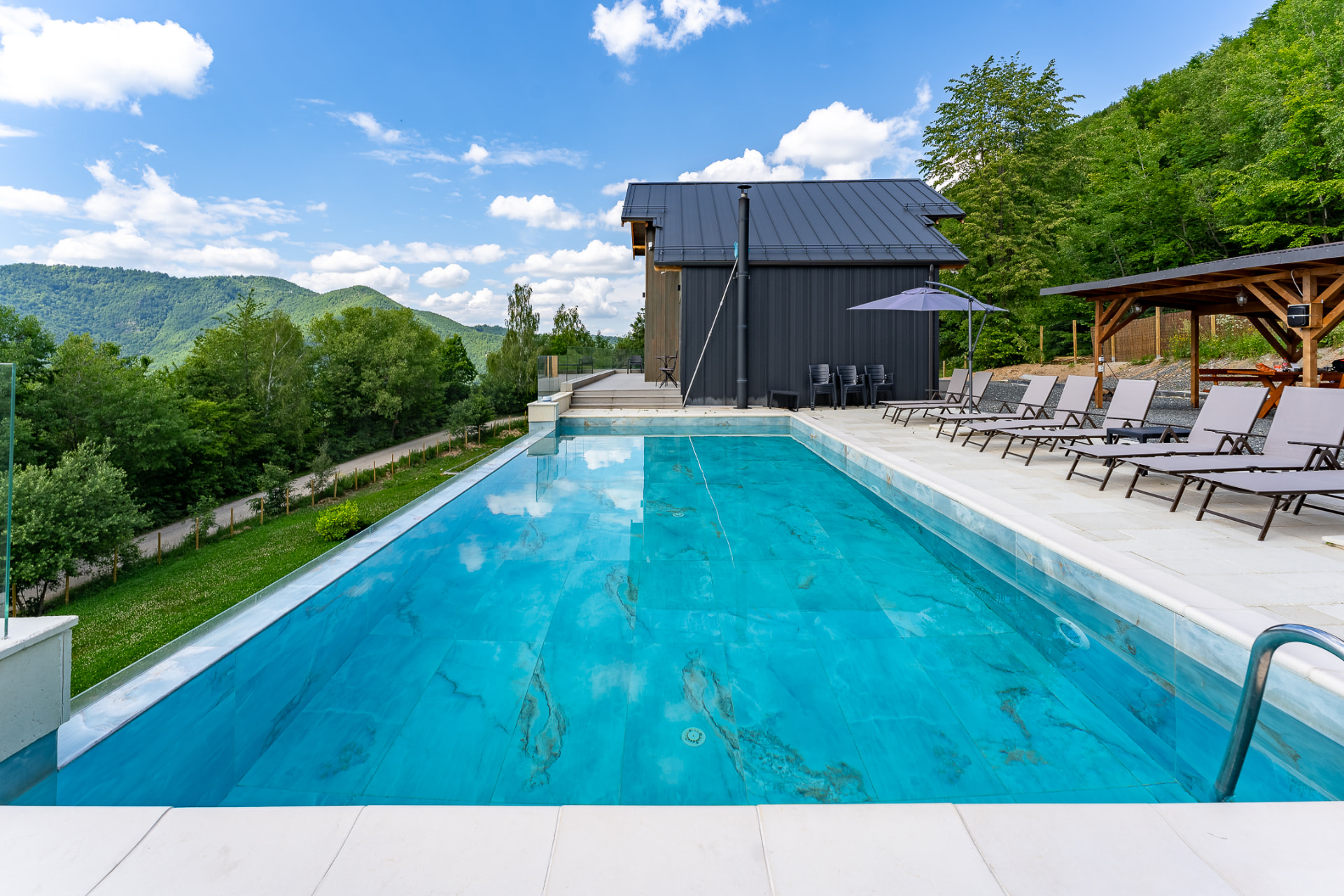 Piscina lungă cu apă turcoaz, cabana cu fațadă neagră și foișor de lemn, înconjurate de pădure